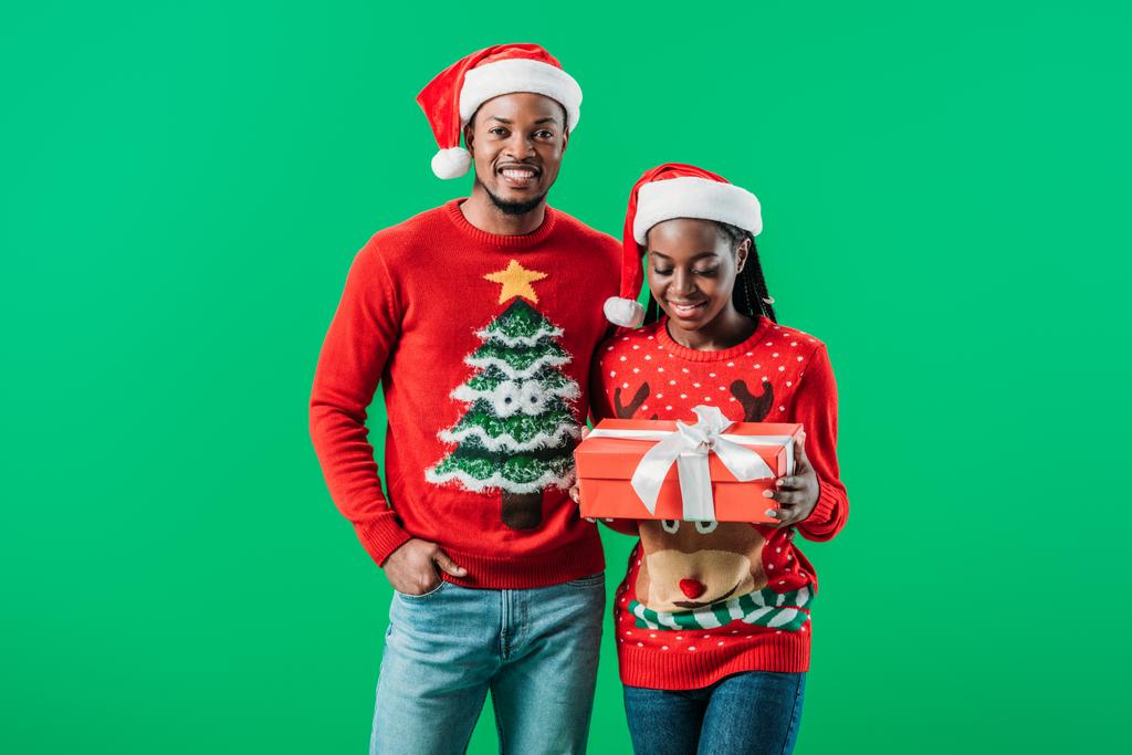 赤いクリスマスセーターとサンタ帽子のアフリカ系アメリカ人男性は、緑の上に手で隔離されたギフトボックスを保持している女性の近くに - 写真・画像