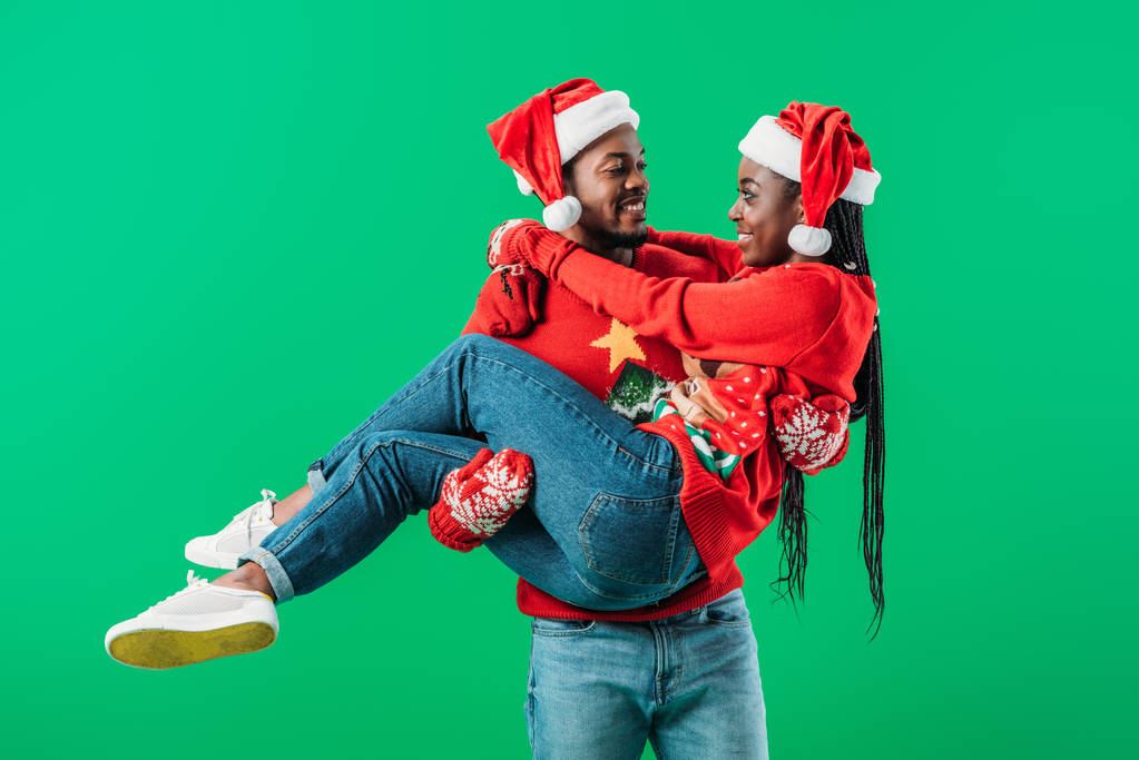 African American man in red Christmas sweater and Santa hat holding up smiled woman isolated on green - Photo, Image