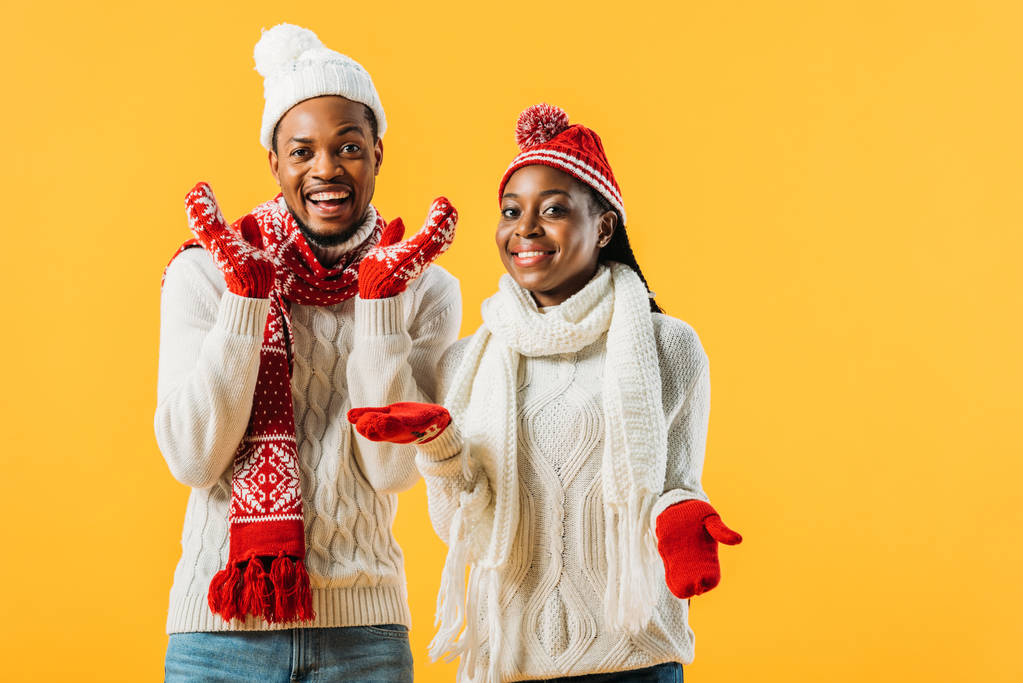 ニットセーター、スカーフ、手袋を着たアフリカ系アメリカ人のカップルが微笑み、黄色で隔離されたカメラを見ている - 写真・画像