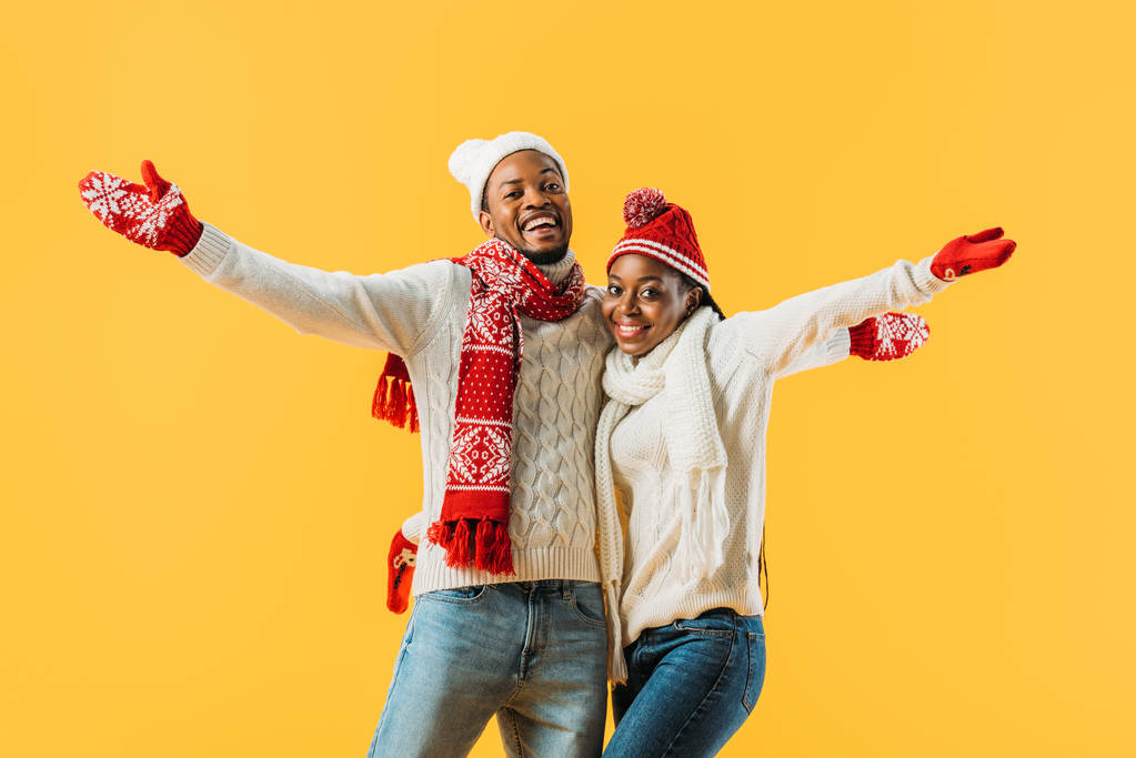 居心地の良い冬の衣装でアフリカ系アメリカ人のカップルは、手を伸ばし、黄色で隔離されたカメラを見て抱きしめる - 写真・画像