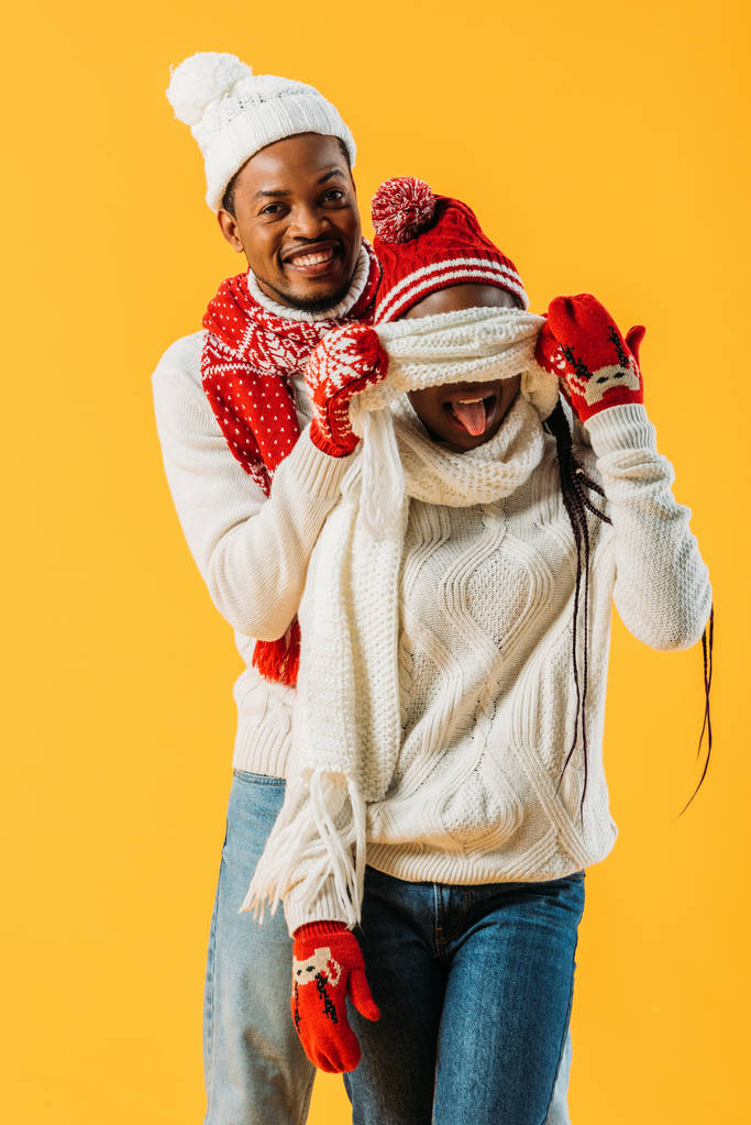 African American man in cozy winter outfit covering with scarf eyes of woman who sticking out tongue isolated on yellow - Photo, Image