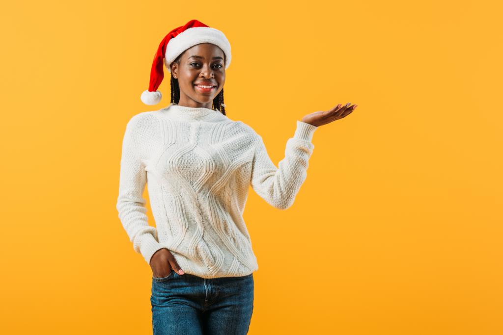 African American woman in winter sweater and Santa hat hold hand in pocket isolated on yellow - Photo, Image