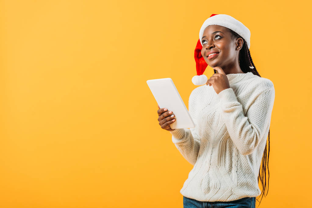dreamy African American woman in winter sweater and Santa hat holding digital tablet isolated on yellow - Fotografie, Obrázek