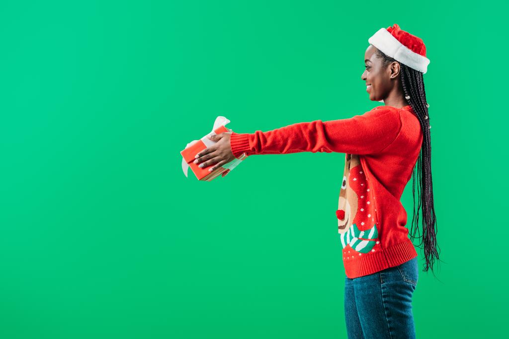 side view of African American young woman in Christmas sweater and Santa hat raising hands with gift box isolated on green - Photo, Image