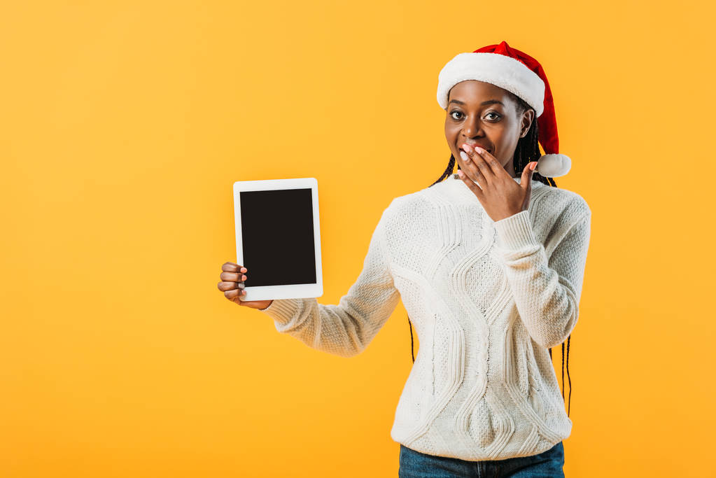 afrikanisch-amerikanische Frau in Winterpullover und Weihnachtsmannmütze mit digitalem Tablet mit leerem Bildschirm und Mund mit Hand isoliert auf gelb - Foto, Bild