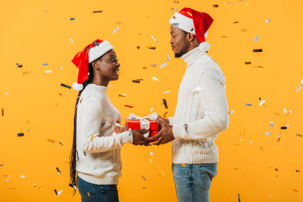 side view of African American couple in Santa hats holding gift box and looking at each other on yellow background with confetti - Fotoğraf, Görsel