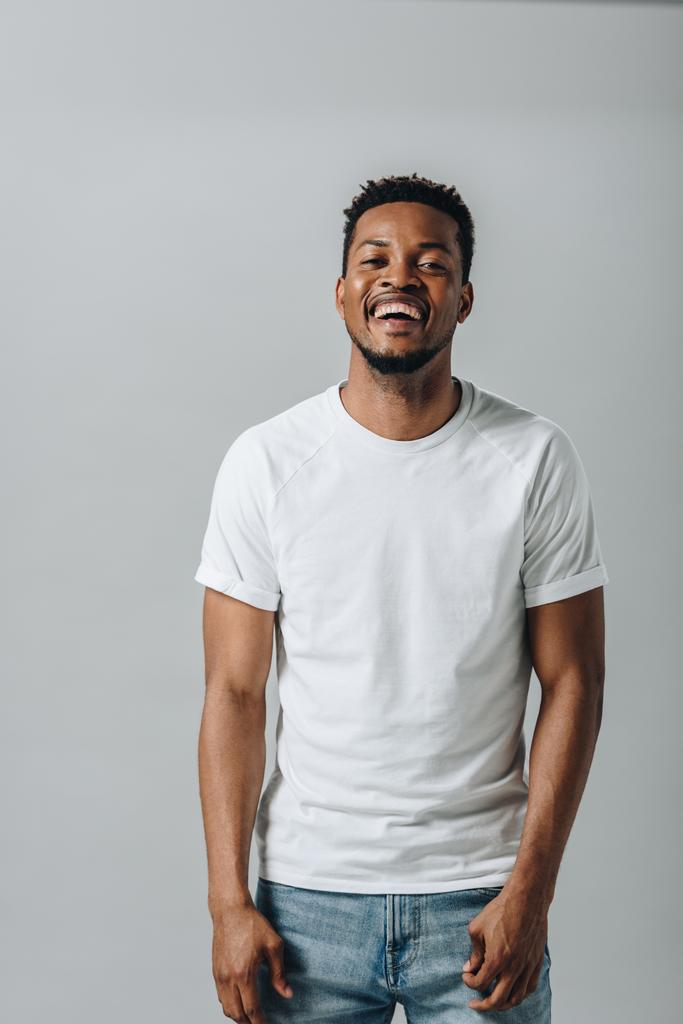 Uomo afroamericano in bianco T-short ridere e guardando la fotocamera isolata su grigio
 - Foto, immagini