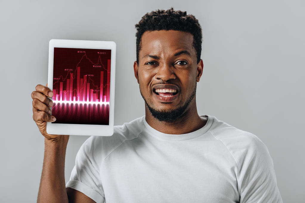 upset African American man holding digital tablet with business charts and looking at camera isolated on grey - 写真・画像