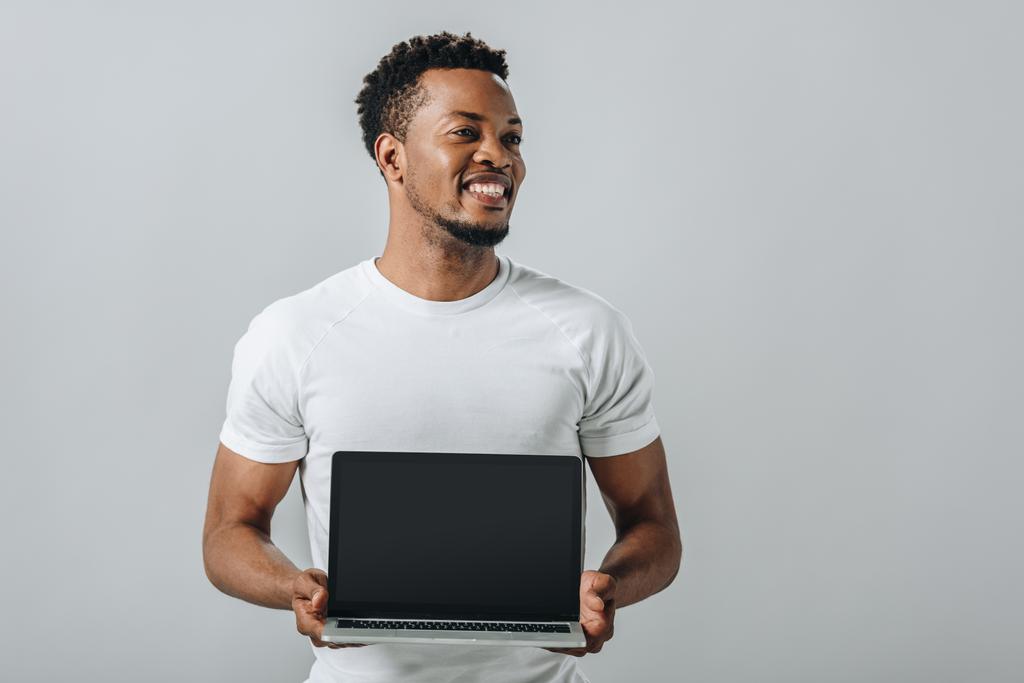homem afro-americano sorrindo e mostrando laptop com tela em branco e olhando para longe isolado em cinza
 - Foto, Imagem
