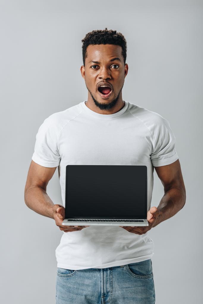 shocked African American man showing laptop with blank screen and looking at camera isolated on grey - Photo, Image