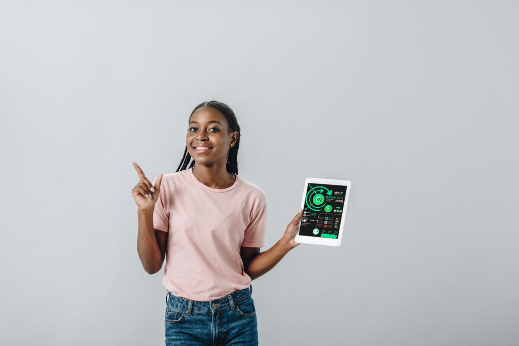 Mulher afro-americana segurando tablet digital com infográficos e apontando com o dedo isolado em cinza
 - Foto, Imagem