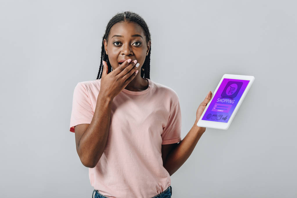 Афроамериканка держит цифровой планшет с приложением для покупок и закрывает рот рукой, изолированной на сером
 - Фото, изображение