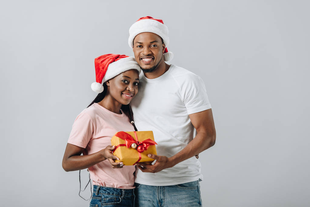 African American couple in Santa hat embracing and holding gift box isolated on grey - Photo, Image