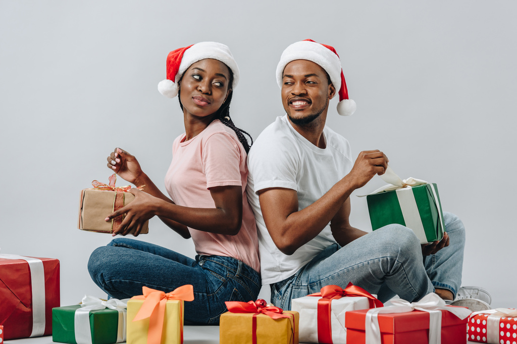 Αφρικανικό ζευγάρι με καπέλο Σάντα που κάθεται πίσω στην πλάτη, ανοίγοντας δώρα και κοιτάζοντας το ένα το άλλο απομονωμένο σε γκρι - Φωτογραφία, εικόνα