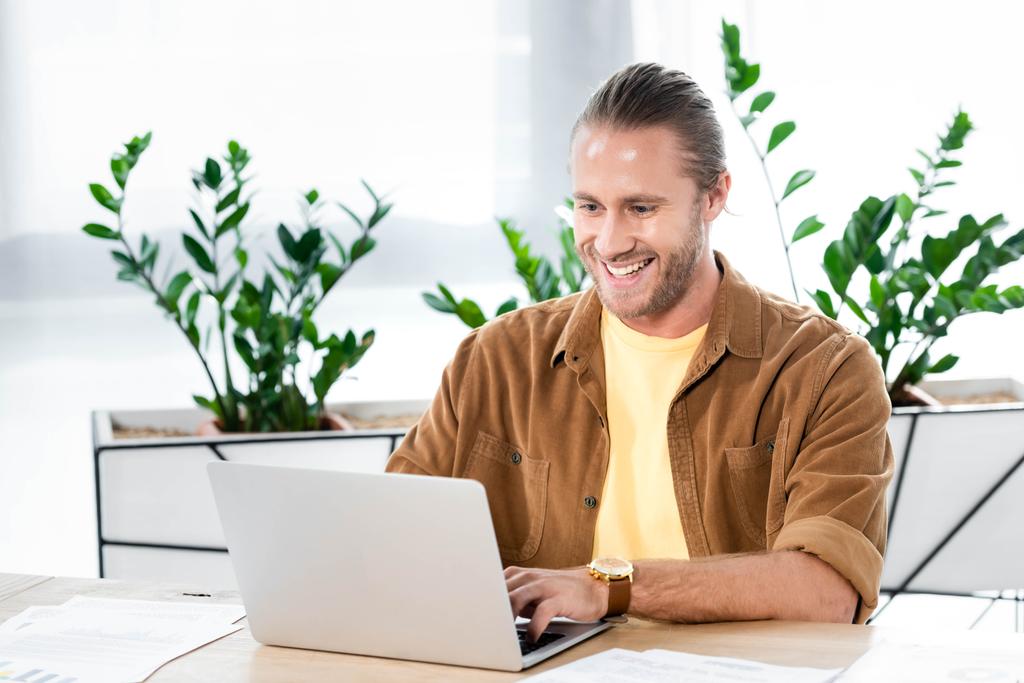 όμορφος και χαμογελαστός επιχειρηματίας κάθεται στο τραπέζι και χρησιμοποιώντας το laptop στο γραφείο  - Φωτογραφία, εικόνα