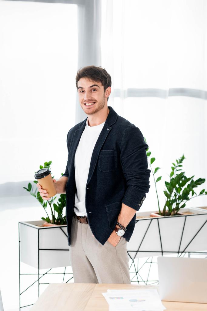 pohledný a usměvavý muž přidržjící papírový pohár a dívat se na kameru v kanceláři  - Fotografie, Obrázek