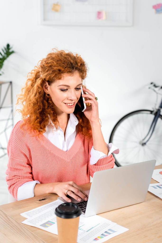 atractiva mujer de negocios en suéter rosa hablando en el teléfono inteligente y utilizando el ordenador portátil en la oficina
  - Foto, Imagen