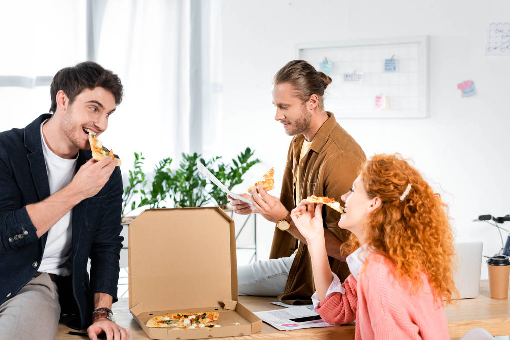 três amigos sorridentes comendo pizza e fazendo papelada no escritório
  - Foto, Imagem