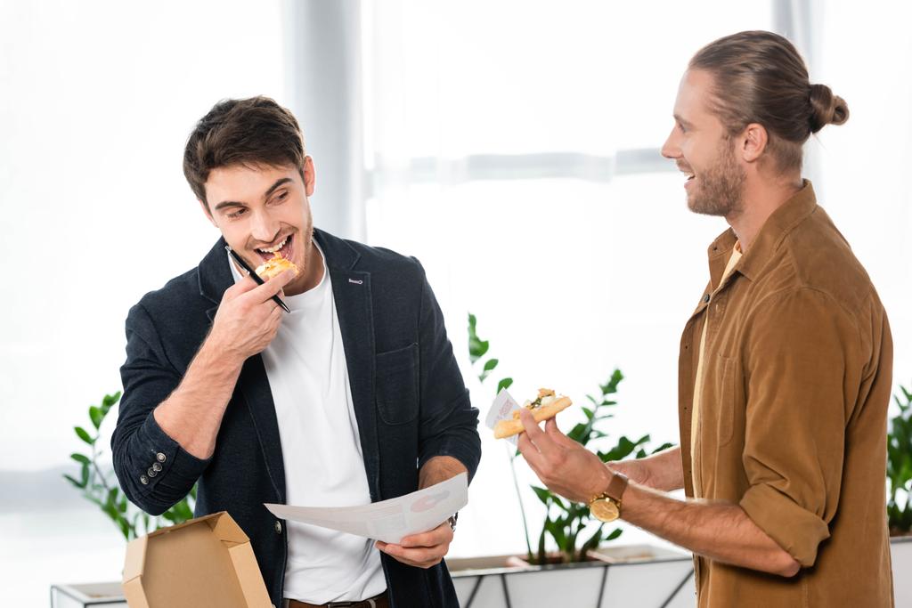 ピザを食べ、オフィスで紙を持っている2人の笑顔の友人  - 写真・画像
