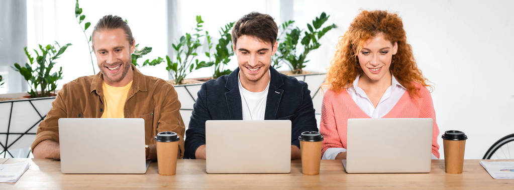 Panoramaaufnahme von drei lächelnden Freunden mit Laptops im Büro  - Foto, Bild