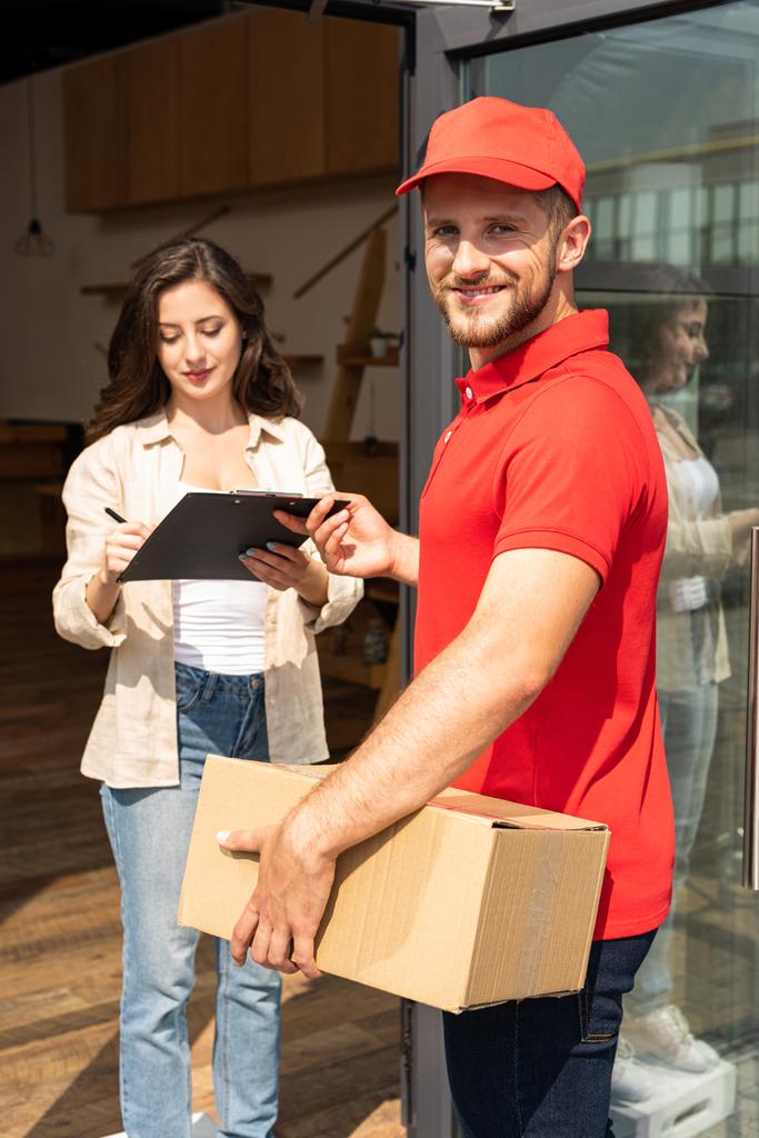 happy delivery man with box holding clipboard near attractive girl  - Photo, Image