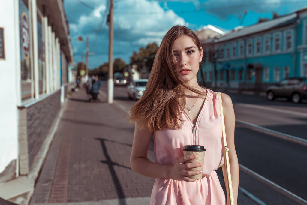 Menina bonita em vestido rosa, fundo de nuvens de construção de estradas, segurando uma xícara de chá de café. No verão na cidade, esperando por namoradas e amigos, cabelo magnífico, espaço livre para texto
. - Foto, Imagem
