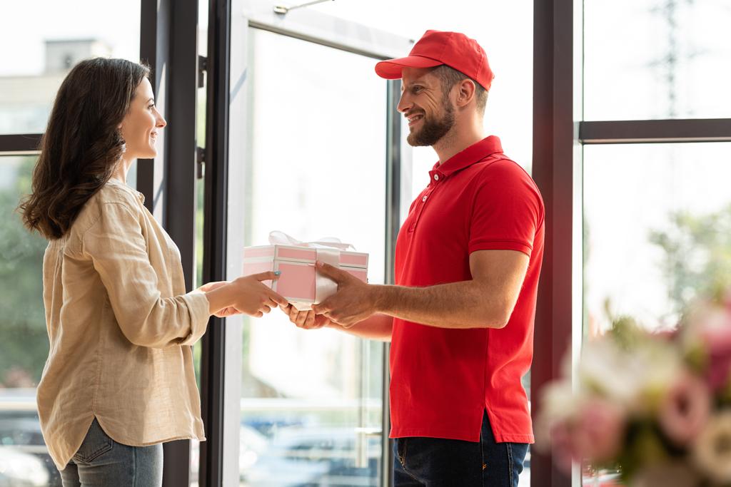 selective focus of handsome delivery man in cap giving pink present to happy woman  - Photo, Image