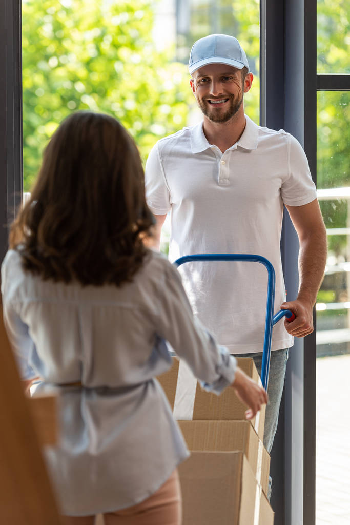selective focus of happy delivery man holding delivery cart with boxes near woman  - Photo, Image