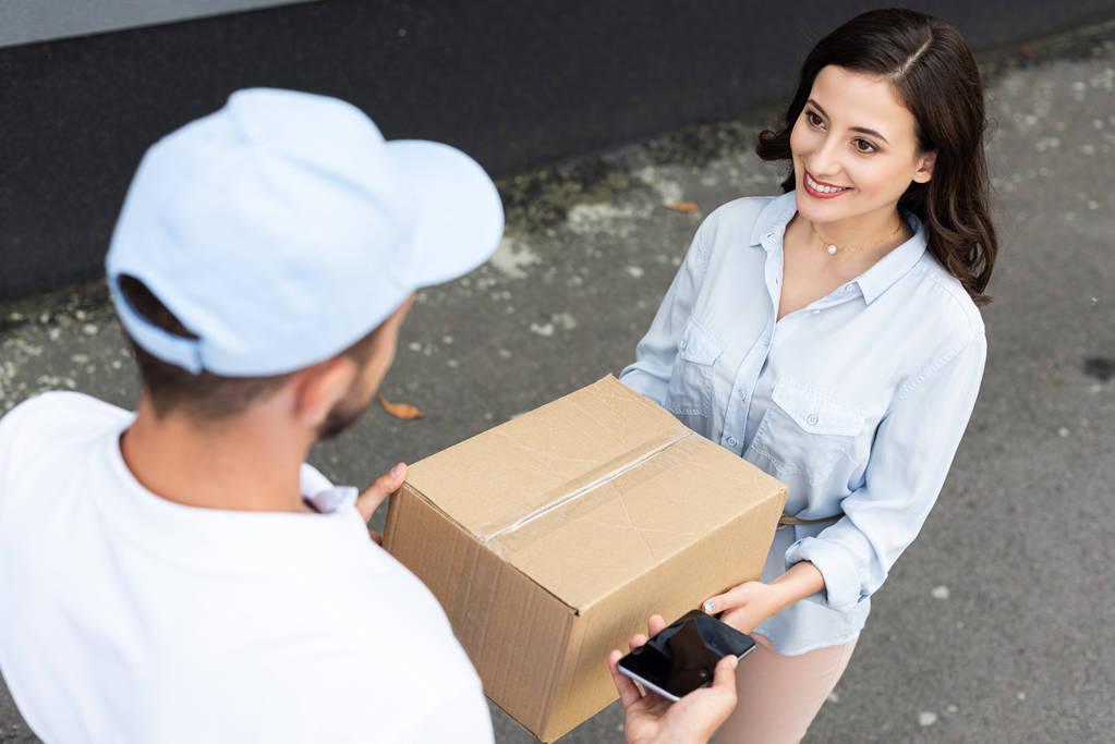 bovenaanzicht van de levering man in Cap geven box aan gelukkige vrouw terwijl het nemen van een smartphone met blanco scherm buiten  - Foto, afbeelding
