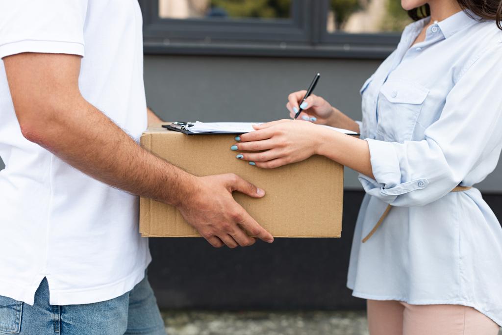cropped view of man holding box near woman signing paper on clipboard outside  - Photo, Image