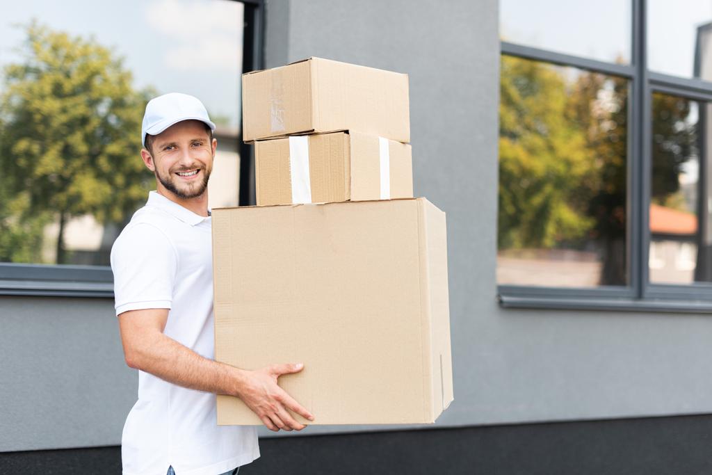 cheerful delivery man in cap looking at camera and holding boxes  - Photo, Image