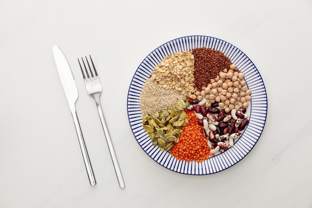 bovenaanzicht van de vork en het mes in de buurt van gestreepte plaat met rauwe linzen, kikkererwten, quinoa, havermout, bonen en pompoenpitten op marmeren oppervlak - Foto, afbeelding