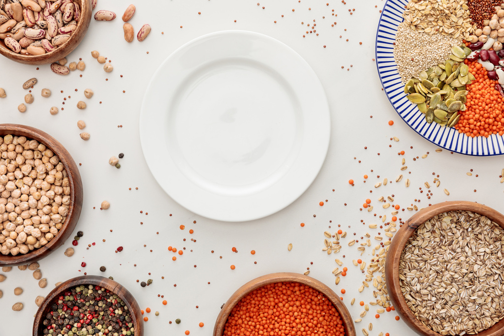 Draufsicht auf leere Teller und Schalen mit rohen Linsen, Quinoa, Haferflocken, Bohnen, Pfefferkörnern und Kürbiskernen auf Marmoroberfläche mit verstreuten Körnern - Foto, Bild