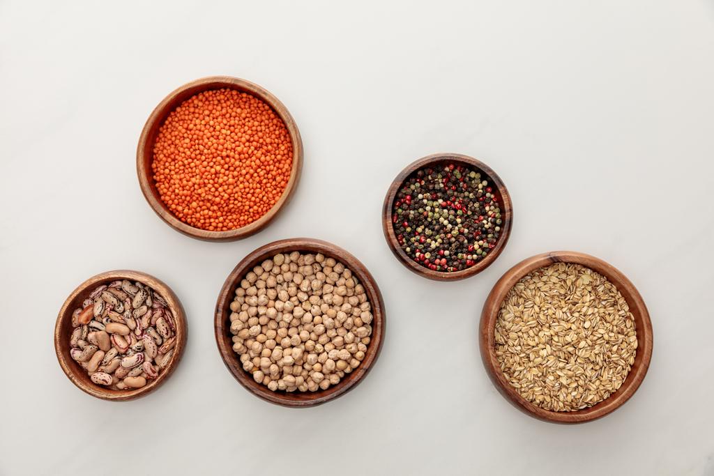 top view of wooden bowls with red lentil, beans, chickpea, oatmeal and peppercorns on white marble surface - Photo, Image