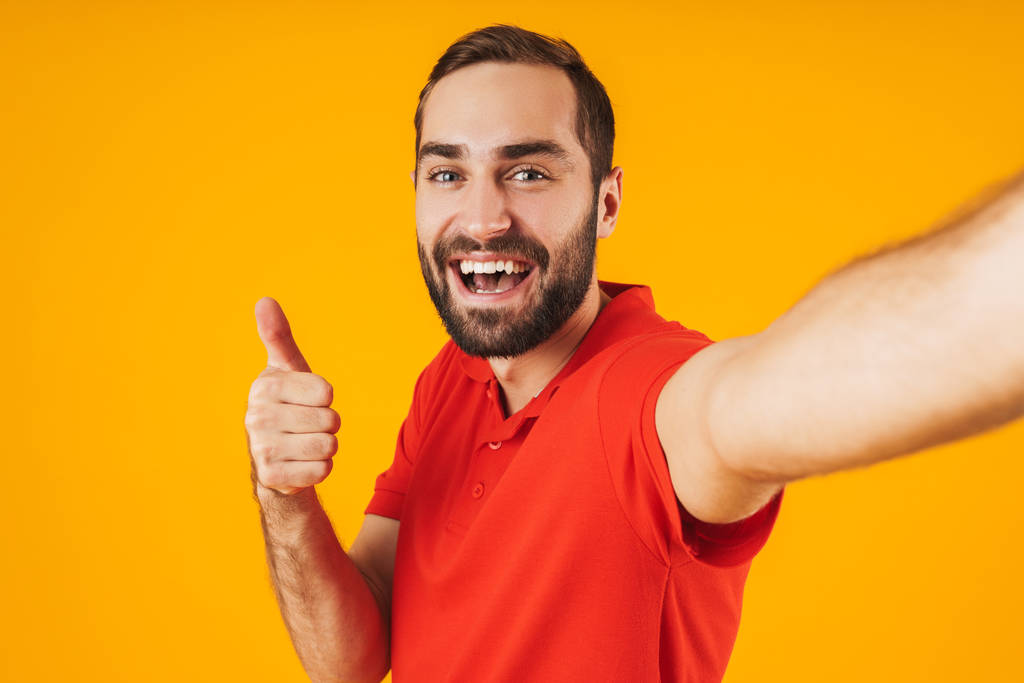 Portrét potěšené muže, který se smál a ukazoval na palec, když si vzal fotografii izolovanou přes žluté pozadí - Fotografie, Obrázek