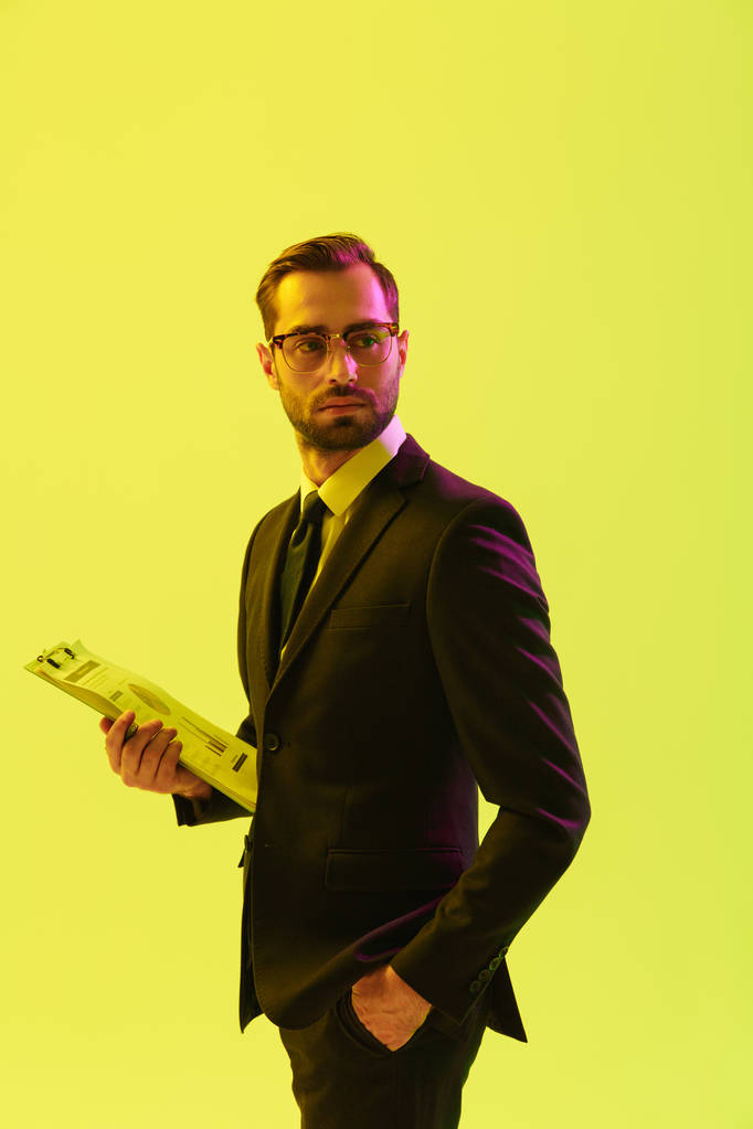 attraktive konzentrierte gutaussehende junge Geschäftsmann posiert isoliert über hellgrünem Hintergrund Wand mit LED-Neonlichtern halten Klemmbrett Schreibnotizen. - Foto, Bild