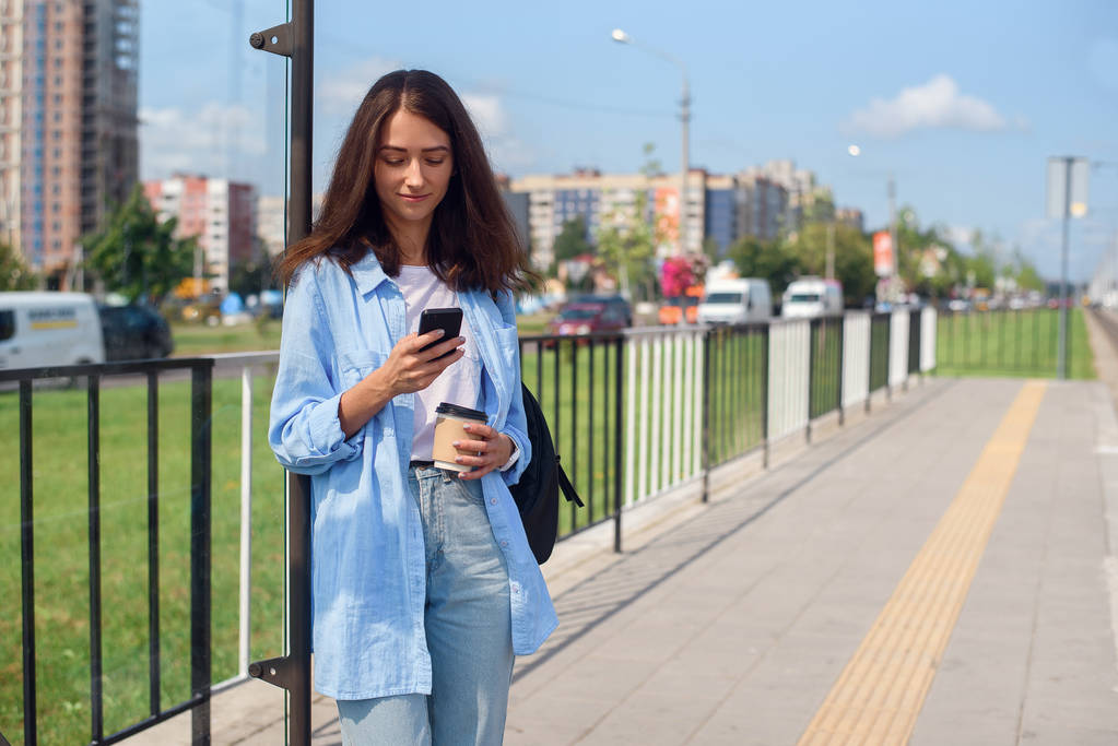 コーヒーを飲んだかわいい女の子は、午前中に公共交通機関の駅でバスやトラムを待っています。アプリを介してスマートフォンの監視輸送を持つ若い女性. - 写真・画像