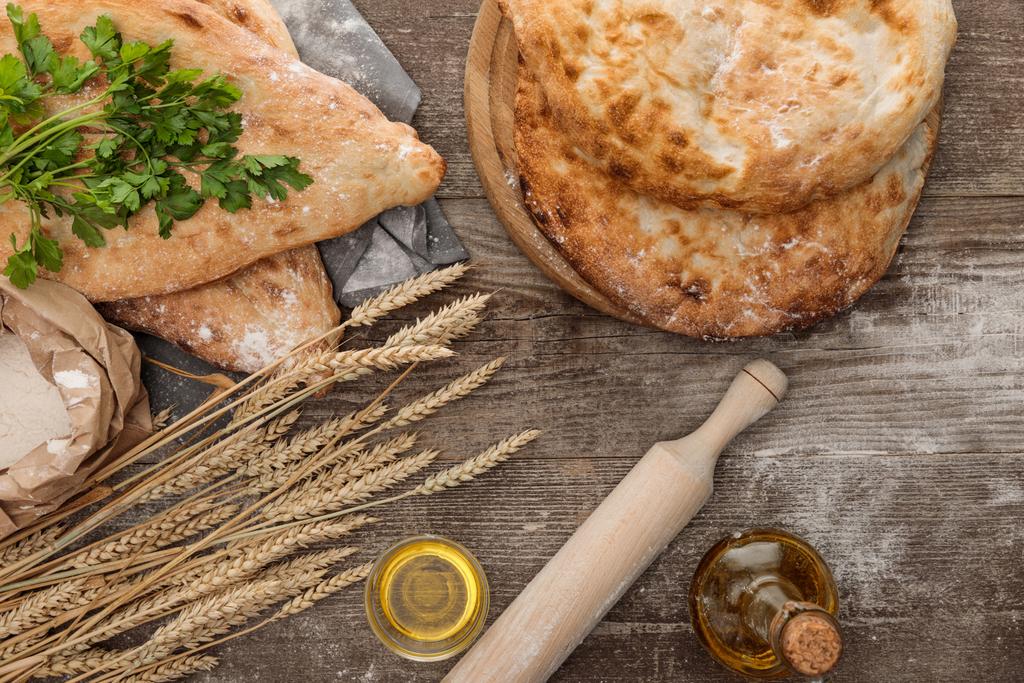 top view of lavash bread on gray towel near flour package, wheat spikes, fresh greenery and olive oil on wooden table - Photo, Image