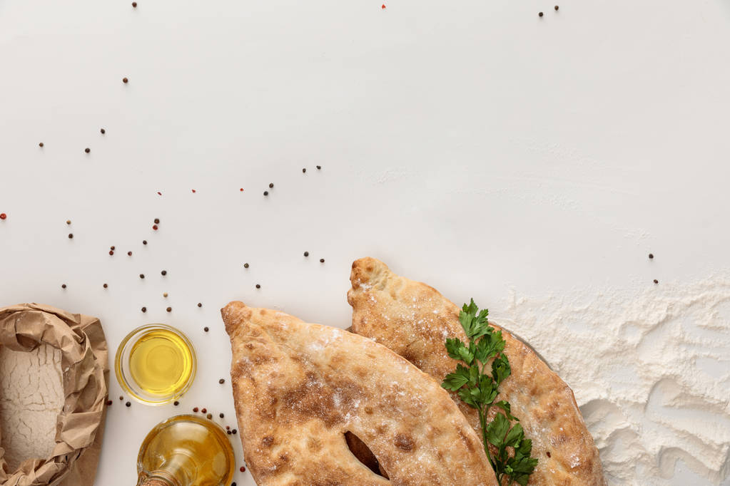 Κορυφαία όψη του ψωμιού lavash με φρέσκο μαϊντανό κοντά στο αλεύρι και το ελαιόλαδο σε λευκή επιφάνεια με κόκκους πιπεριού - Φωτογραφία, εικόνα