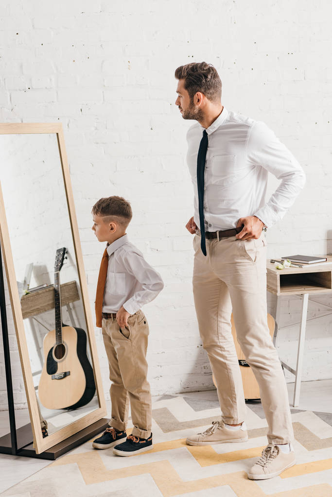 volledige lengte weergave van zoon en vader in formele slijtage staande in de buurt van spiegel - Foto, afbeelding