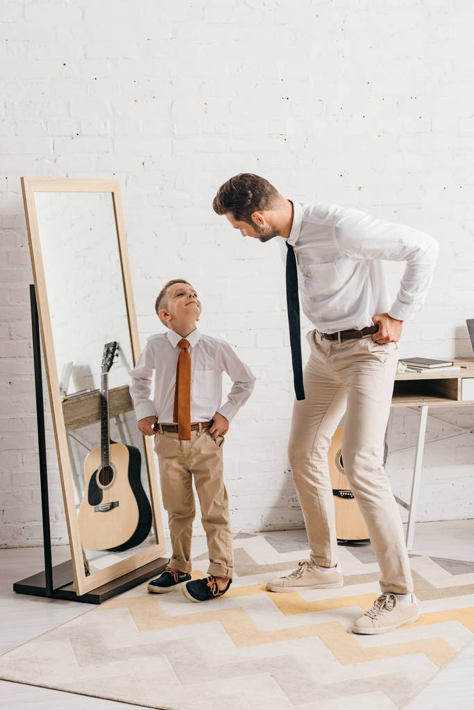 Ganzkörperansicht von Sohn und Vater in formeller Kleidung, die in der Nähe des Spiegels stehen und einander anschauen - Foto, Bild