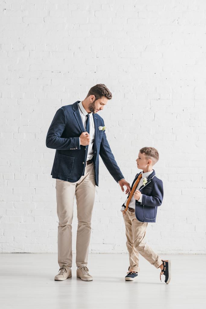 Ganzheitliche Ansicht von Vater und Sohn in Jacken mit Boutonnieres, die sich gegenseitig anschauen - Foto, Bild