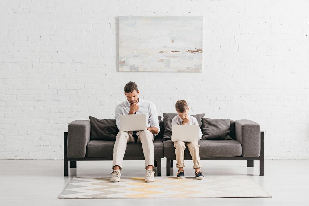 padre e hijo en ropa formal usando computadoras portátiles mientras están sentados en el sofá
 - Foto, imagen
