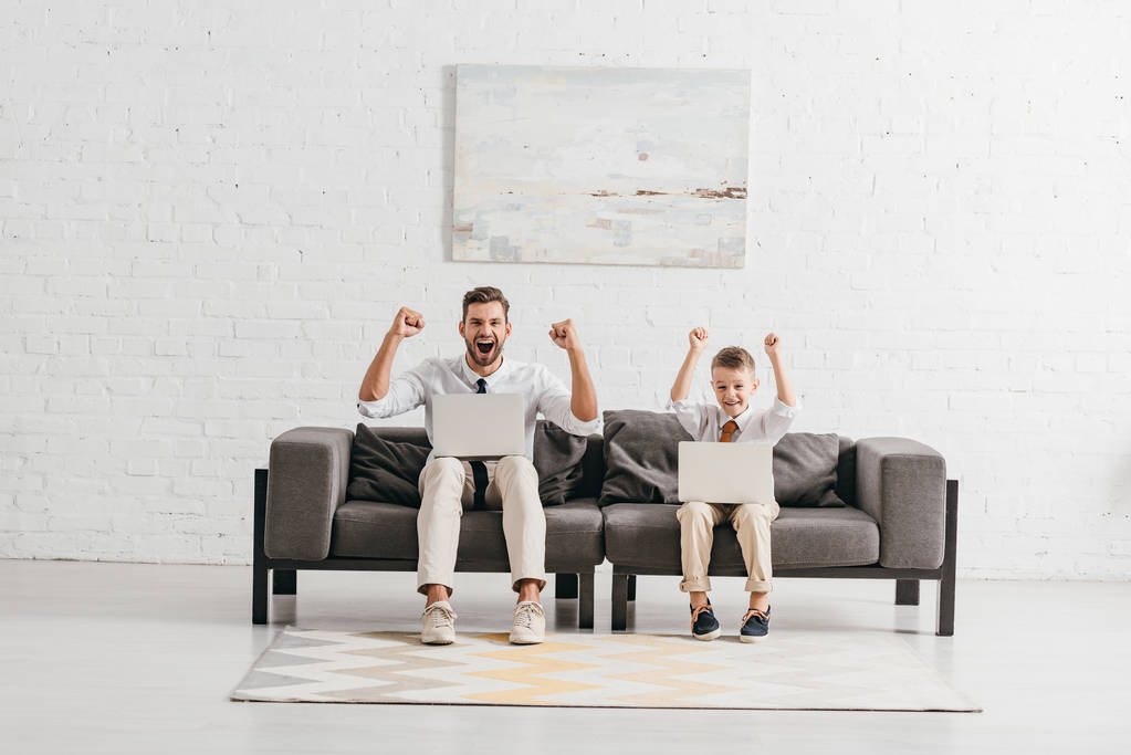 pai e filho em desgaste formal usando laptops e mostrando sim gestos enquanto sentado no sofá
 - Foto, Imagem