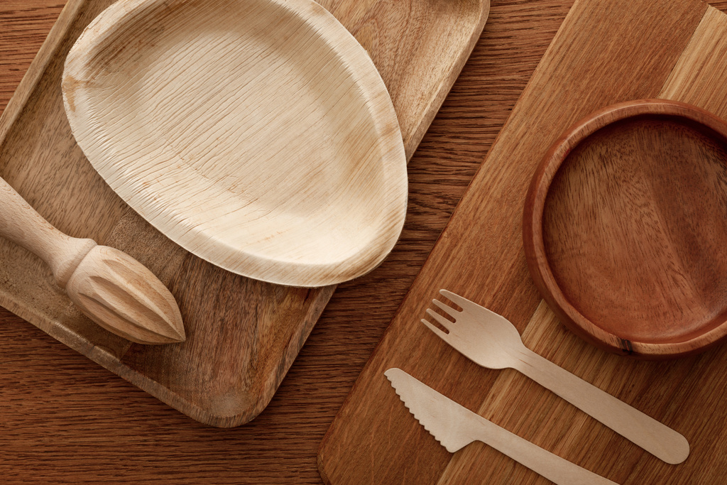 верхний вид деревянной посуды и доски для резки с тарелками, столовыми приборами и соковыжималкой для рук на коричневом фоне
 - Фото, изображение