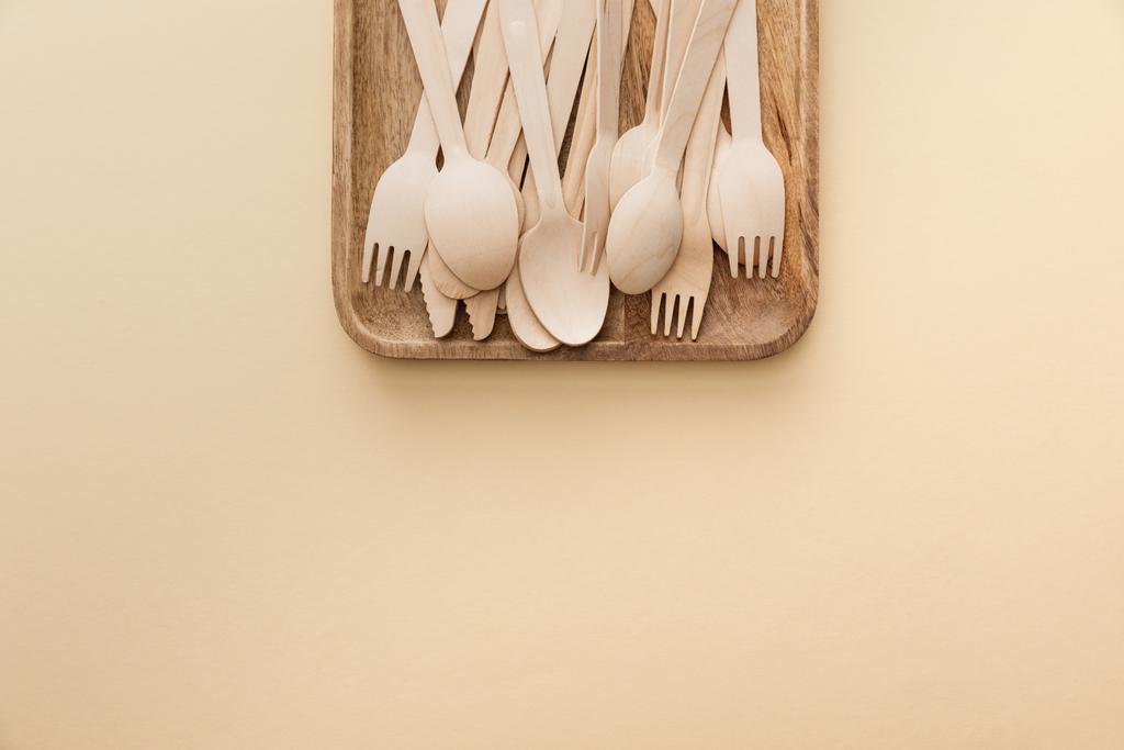 pohled na pravoúhlý dřevěný talíř s vidlemi, nožem a lžičky na béžové pozadí - Fotografie, Obrázek