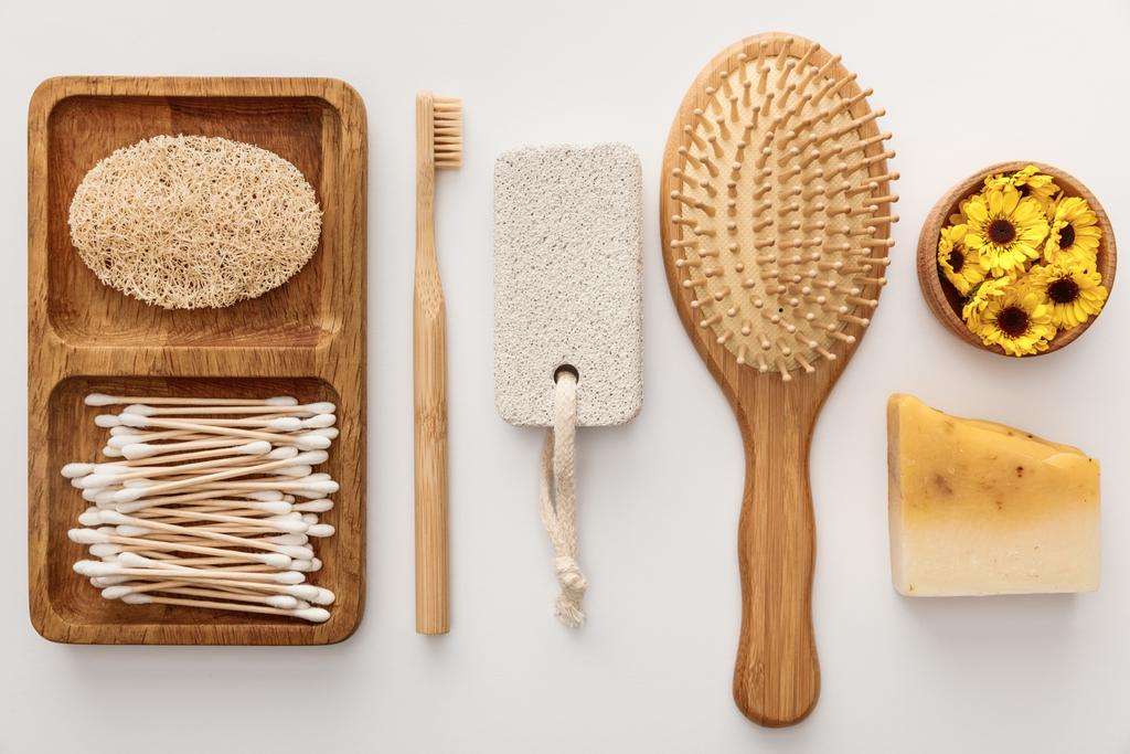 flat lay with wooden dish with cotton swabs and loofah near toothbrush, hairbrush, piece of soap, pumice stone and cup with flowers on white background - Photo, Image