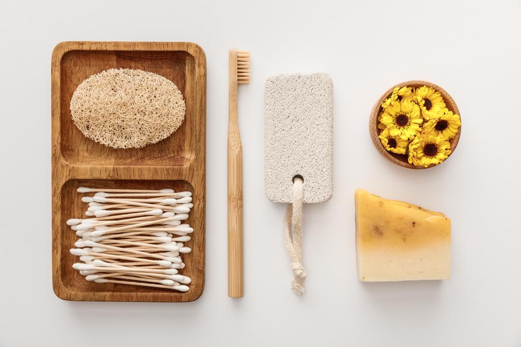 platte lay met houten schotel met wattenstaafjes en Loofah in de buurt van tandenborstel, stuk van zeep, puimsteen en een kopje bloemen op witte achtergrond - Foto, afbeelding