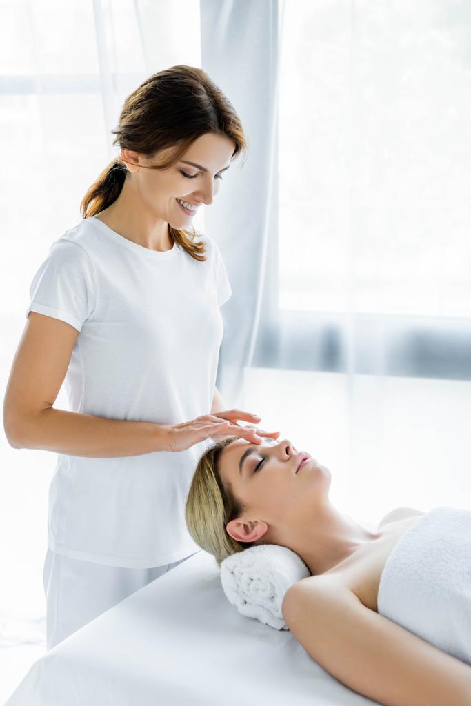 massagista sorrindo fazendo massagem facial para mulher atraente no spa
  - Foto, Imagem