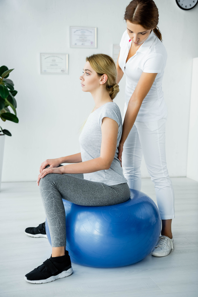 attraktive Patientin sitzt auf blauem Gymnastikball und Chiropraktiker berührt ihren Rücken  - Foto, Bild